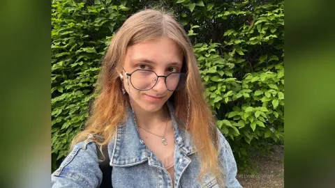 Young woman with long blonde hair and glasses wearing a denim jacket in front of lush green foliage.