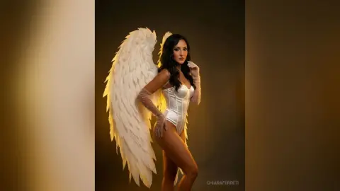 Elegant woman in white corset and large angel wings posing in a studio with warm lighting.