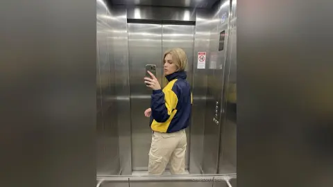 CharlotteReimer takes a mirror selfie in an elevator wearing a blue and yellow jacket and khaki cargo pants.
