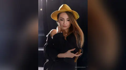 Elegant woman in a black outfit and yellow wide-brimmed hat posing in moody, cinematic lighting.