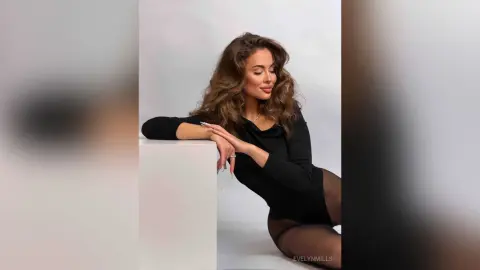 EvelynMills poses leaning against a white block wearing a black long-sleeve bodysuit and sheer black tights.