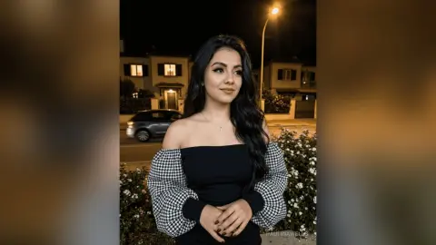 Young woman in a black off-the-shoulder top and checkered sleeves posing outdoors at night under streetlights.