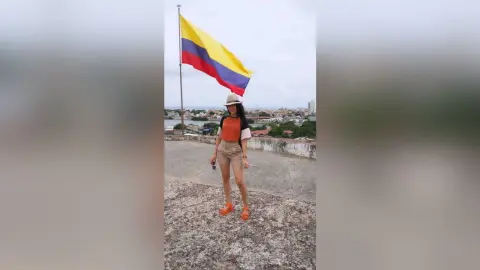 JessieLiu stands outdoors wearing a white hat, an orange tank top, and tan shorts against a background of a large yellow, blue, and red flag.