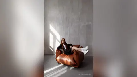 Redheaded woman in a black blazer and white trousers relaxing on a brown leather sofa in a minimalist room.