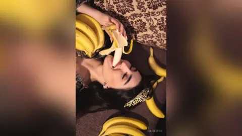 JuliDeming lies on a patterned surface with long dark hair, playfully biting into a peeled banana surrounded by several more bananas.