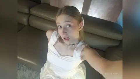 EvelinaMoorina wearing a white dress while sitting on a grey couch and looking into the camera.