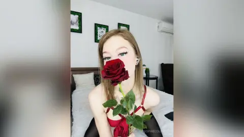 Blonde woman in a red lace bodysuit holding a single red rose in a bright bedroom setting.