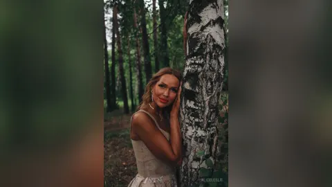 AliceLeslie — AliceLeslie leans against a birch tree in a forest wearing a beige sleeveless top.