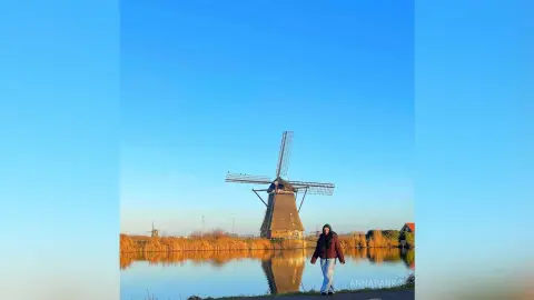 AnnaBanks stands by a calm lake in front of a large windmill under a clear blue sky, wearing a brown jacket and white pants.