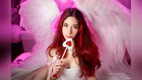 Red-haired angel cosplay in white dress with large feathered wings holding a heart wand in soft pink lighting.