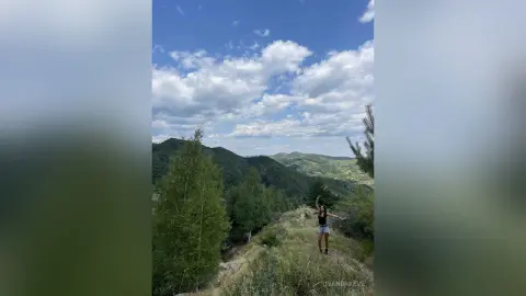 DyandraEve stands on a grassy mountain ridge overlooking a vast, green forest valley under a blue sky with white clouds.