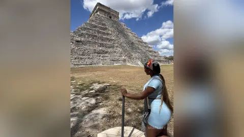 Woman in a blue summer outfit standing before the ancient Chichen Itza Mayan pyramid under a sunny sky.