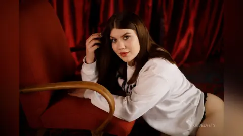 NikkiVelvets — Young woman in a white sweatshirt relaxing on a red velvet chair against a moody red curtain background.