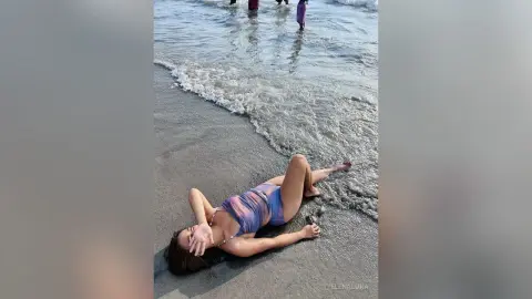 Young woman in a purple patterned swimsuit relaxing on a sandy beach by the ocean waves.