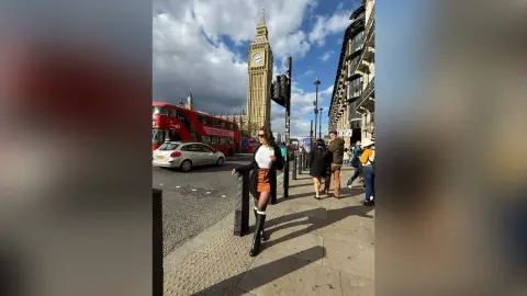 Stylish woman in a white top and orange skirt walking near Big Ben and a red London bus on a sunny day.
