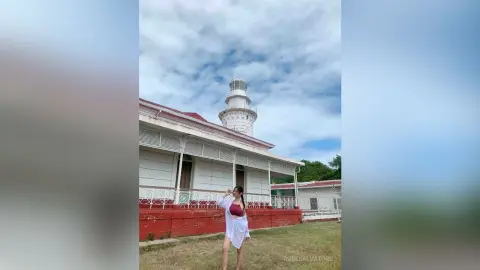 Young woman in a white summer outfit standing before a historic white lighthouse under a cloudy blue sky.