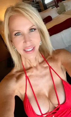 Smiling blonde woman in a red bikini top posing in a bright bedroom setting.