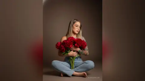 Young woman with glasses in denim jeans holding a large bouquet of red flowers in a moody studio setting.