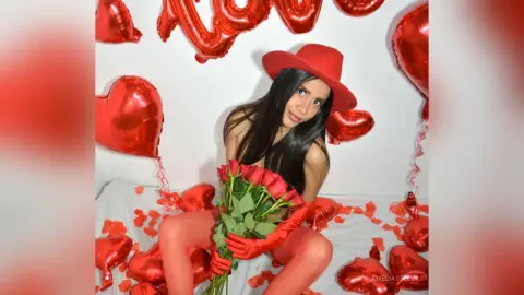 AmeliaStell poses naked with a bouquet of red roses, wearing a red hat and red gloves against a white background with red heart balloons.