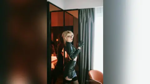 Stylish blonde woman in a black leather outfit and glasses posing in a modern room with moody lighting.