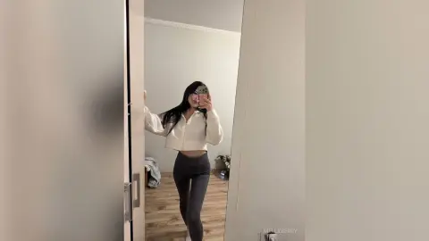 Young woman in a white cropped sweatshirt and grey leggings taking a mirror selfie in a bright minimalist room.
