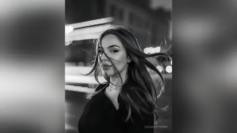 Moody black and white portrait of a woman with long flowing hair in a dark outfit against blurred city lights.