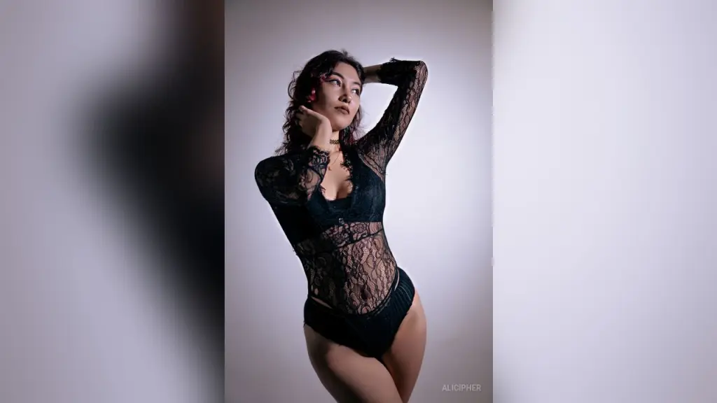 Elegant woman in a black sheer mesh bodysuit sitting on a red velvet sofa in a moody, dimly lit room.