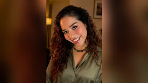 Smiling young woman with curly hair wearing an olive green blouse in a warm indoor setting.