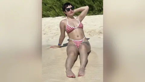 Woman in red checkered bikini relaxing on a sunny sandy beach under bright sunlight.