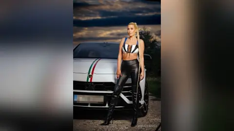 Confident blonde woman in black leather pants and white crop top posing by a white car at sunset.