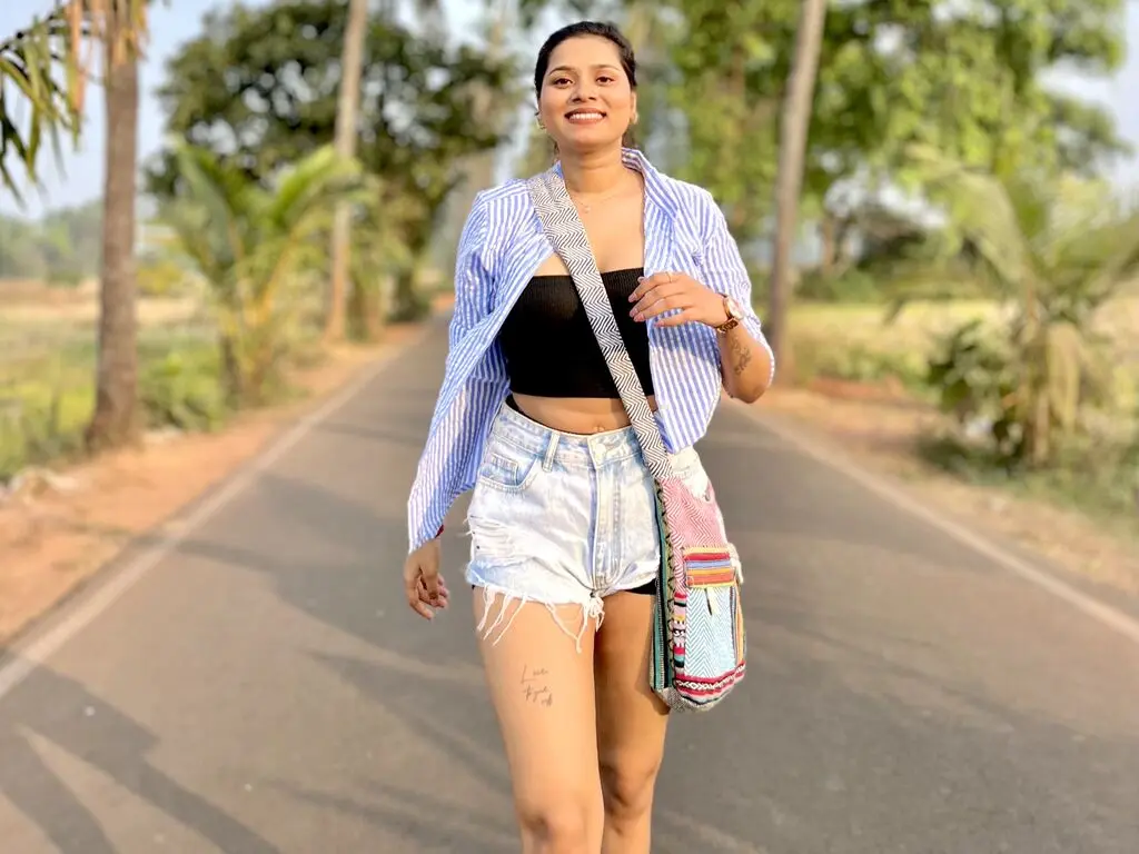AnnaIver smiles while wearing a black tube top, light denim shorts, and a blue striped button-down shirt, carrying a colorful woven bag on a paved road.