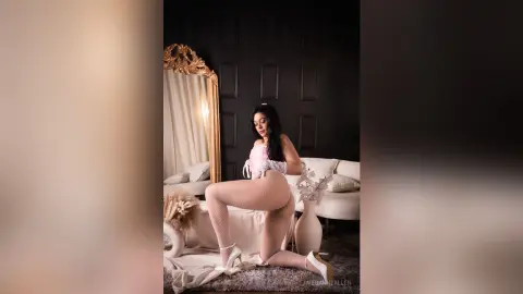Elegant woman in pink lace lingerie and fishnet tights posing in a moody, luxury white bedroom setting.