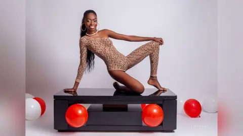 Smiling Black woman in a leopard print bodysuit posing on a black platform with red and white balloons.