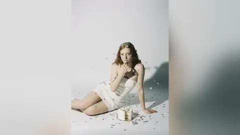 Redheaded woman in a white dress sitting on a studio floor with colorful confetti for a celebratory mood.