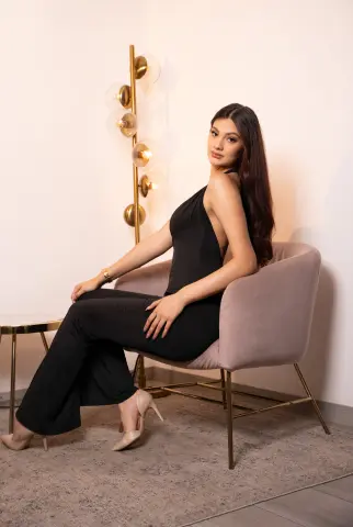Elegant woman in a black jumpsuit sitting in a modern chair against a minimalist interior background.