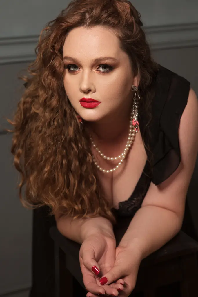 SinFulTitsDD with red hair poses behind a table of fruit, wearing a black mesh top and large green earrings.