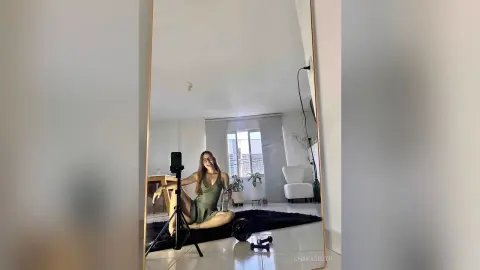 Young woman in a green satin dress taking a mirror selfie in a bright, modern minimalist living room.