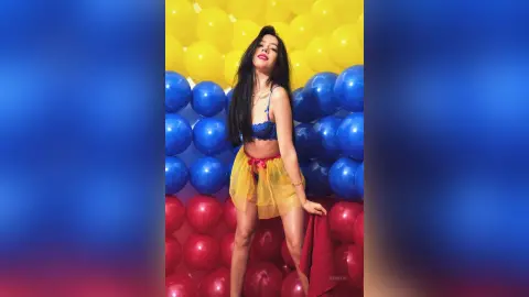 JessieLiu poses in a blue lace bra and a sheer yellow skirt against a background of yellow, blue, and red balloons.