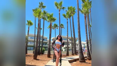 DesyreeDawson stands outdoors in a white bikini and black jacket, posing between tall palm trees under a clear blue sky.