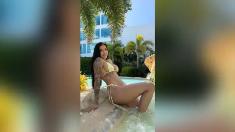 Aleja poses in a yellow string bikini while sitting on a rock near a pool surrounded by palm trees.