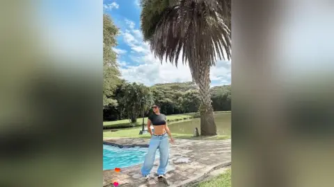 Stylish woman in black crop top and baggy jeans relaxing by a tropical swimming pool under palm trees.