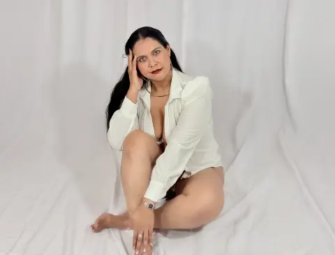 Elegant woman in a white button-down shirt sitting on a white fabric backdrop with a serene expression.