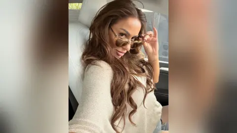 Smiling brunette woman in a cream sweater wearing sunglasses while sitting in a car interior.