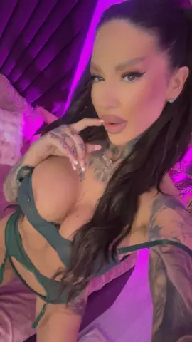Tattooed woman in green lingerie posing in a moody, purple-lit bedroom setting.