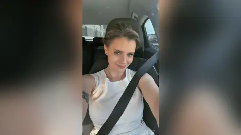 AnishaBee smiles while sitting in a car seat, wearing a white sleeveless top with her hair pulled back.