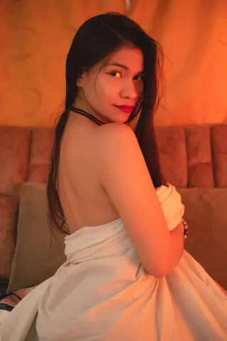Young woman in a white wrap sitting on a bed with warm, moody lighting.