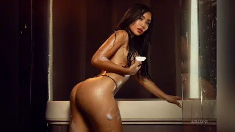 Sensual woman with long dark hair posing in a white bathtub with soap suds in a moody, dimly lit setting.