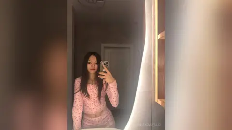 Young woman in a pink floral long-sleeve top taking a moody mirror selfie in a dimly lit room.