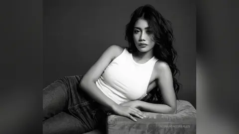 Elegant woman in a white tank top and denim jeans posing gracefully in a moody black and white studio portrait.
