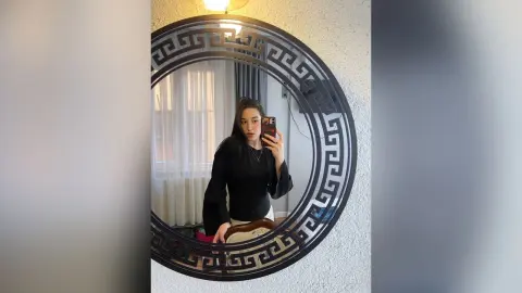 Young woman in a black long-sleeve blouse taking a mirror selfie in a bright, modern room.